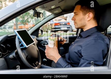 Der Fahrer sieht während der Mittagspause Filme oder Fernsehsendungen auf dem Tablet. Stoppen für einen Happen zu essen. Mann essen Snack im Auto und trinkt Kaffee oder Tee. Stockfoto
