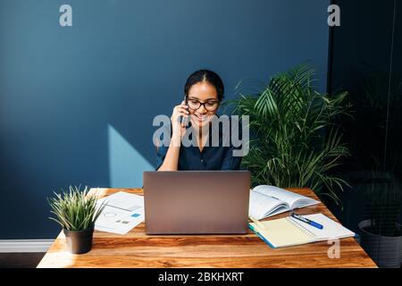 Lächelnde Geschäftsfrau sitzt an einem Tisch und spricht auf dem Handy, während sie am Laptop arbeitet Stockfoto