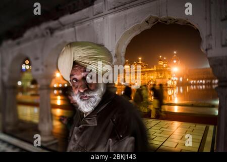 Indien, Punjab Staat, Amritsar, Harmandir Sahib, Porträt eines Sikh-Mannes mit intensiven Augen und Golden Temple beleuchtet in der Nacht im Hintergrund, heiliger Ort des Sikhismus Stockfoto