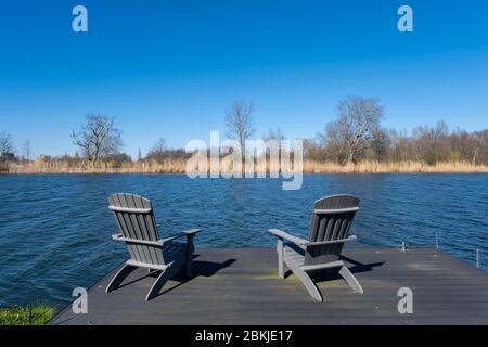 Rustikale handgefertigte Stühle auf einer Holzterrasse mit Blick auf einen Fluss oder See mit Schilf in der Frühlingssonne Stockfoto