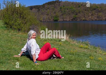 Europa, Luxemburg, Insenborn, attraktive ältere Frau, die während der Covid-19 Lockdown, Frühjahr 2020, am Ufer des Lac Sûre sitzt Stockfoto