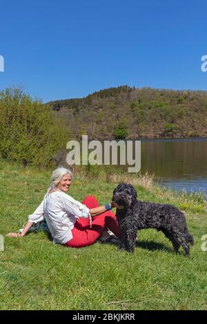 Europa, Luxemburg, Insenborn, attraktive ältere Frau am Ufer des Lac Sûre mit ihrem portugiesischen Wasserhund Stockfoto