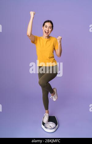 Die volle Länge einer fröhlichen jungen hübschen Sportlerin, die auf der Waage isoliert über violettem Hintergrund steht, feiert Stockfoto