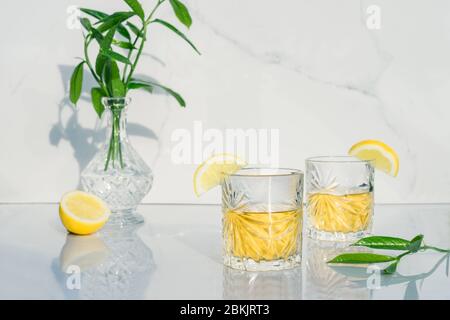 Zwei Kristallgläser mit gelben Cocktails, die mit Zitrone verziert sind. Vase mit frischem Grün und Glas Becher mit grünem Tee, Alkohol auf Marmor Bac Stockfoto
