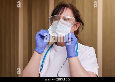 Während der COVID-19-Pandemie legt der Gesundheitsarbeiter eine einmalige Gesichtsmaske an. Kaukasische Ärztin in weißen uniformen Schutzbrille und OP-Handschuhe. Stockfoto