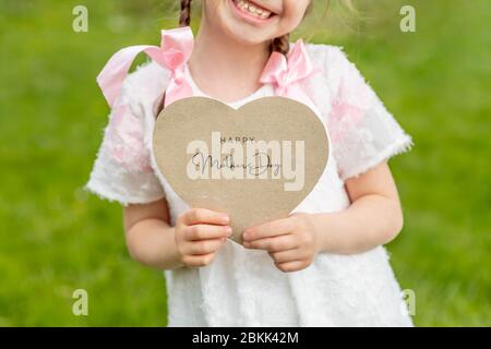 Happy Mother's Day auf einer herzförmigen Karte geschrieben, die von einem Mädchen auf Gras Hintergrund gehalten Stockfoto