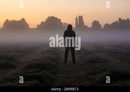 Ein Mann mit Kapuze, der in einem nebligen Feld steht, um in den Himmel zu schauen, kurz vor Sonnenaufgang. Stockfoto