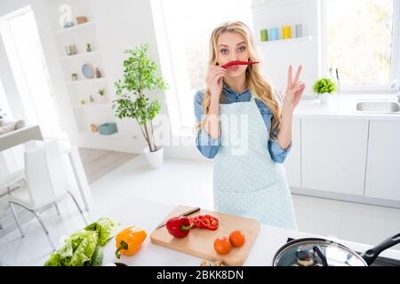 Foto von fröhlichen Hausfrau blonde Dame geschnitten Salat Kochen leckeres Abendessen Frühstück halten rote scharfe Chili Pfeffer wie Schnurrbart zeigt V-Zeichen-Symbol verspielt Stockfoto