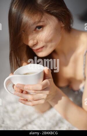 Junge Frau mit einer Tasse Tee in den Händen schaut auf die Kamera. Der Raum ist von Sonnenlicht überflutet Stockfoto