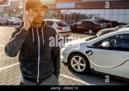 Ein Mann wartet, während sein Elektroauto auf einem Parkplatz in der Nähe eines Einkaufszentrums aufgeladen wird. Stockfoto