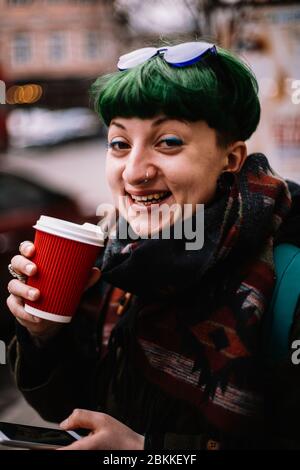 Porträt der glücklichen nicht-binären Hipster Frau Kaffee trinken, während auf der Straße in der Stadt im Winter stehen Stockfoto