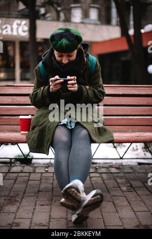 Porträt von glücklichen nicht-binären Hipster mit Smartphone während auf Bank in der Stadt im Winter sitzen Stockfoto