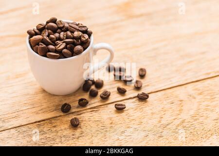 Eine weiße Tasse voll mit braunen Kaffeebohnen auf einem Holztisch Stockfoto