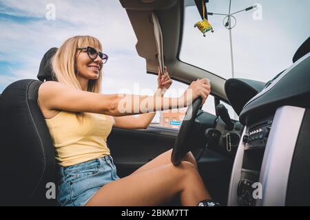 Junge süße Mädchen preens während der Fahrt. Blondine bewundert sich im Spiegel. Glückliche weibliche Fahrer hinter dem Lenkrad. Frau senkt die Sonnenblende hält t Stockfoto