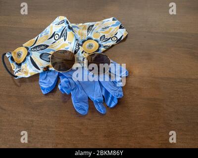 Ein Paar blaue Einweghandschuhe, eine Sonnenbrille und eine handgefertigte Stofffacemaske mit blauem und gelbem Blumenmuster mit weißem Hintergrund auf einer Holzlasche Stockfoto