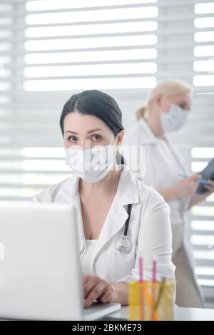 Maskierte Frau mit Stethoskop hinter Laptop, blond am Fenster. Stockfoto