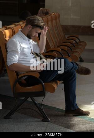 Seitenporträt eines ernsten jungen Mannes, der mit Karte oder Zeitung auf den Zug wartet. Der Reisende sitzt auf der Bank und liest Stockfoto