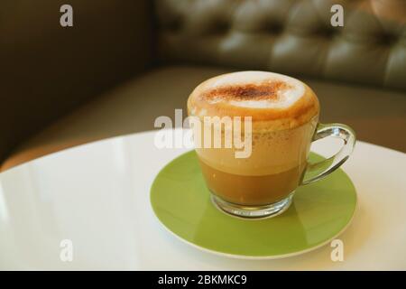 Tasse heißer Cappuccino auf weißem runden Tisch mit unscharfer Couch im Hintergrund serviert Stockfoto