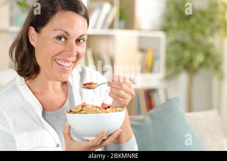 Glückliche Erwachsene Frau mit Müslischale mit Obst beim Frühstück sitzen auf dem Sofa zu Hause Stockfoto