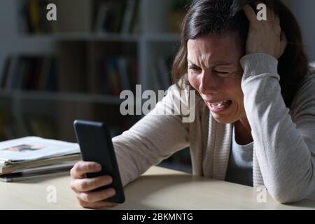 Traurige Erwachsene Frau weint, die schlechte Nachrichten auf dem Smartphone liest und abends im Wohnzimmer zu Hause sitzt Stockfoto