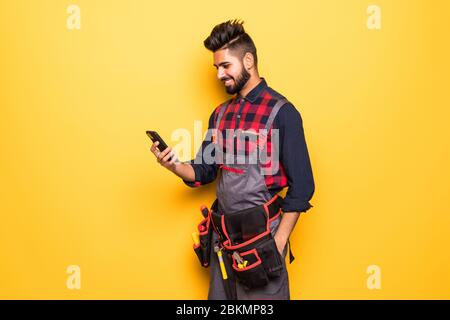 Porträt des jungen Bauarbeiters mit Smartphone in Schutzkleidung, Helm und Werkzeuggürtel isoliert Stockfoto