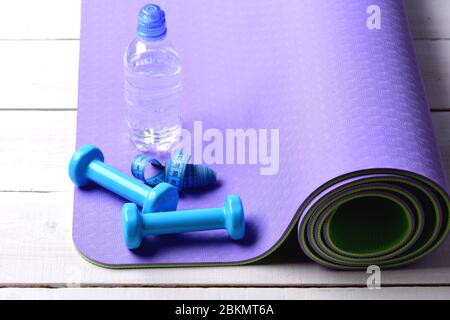 Kurzhanteln aus blauem Kunststoff auf hellem Holzhintergrund. Langhanteln in der Nähe der Wasserflasche und cyanfarbenes Maßband liegen auf lila Yogamatte. Formgebung und Fitnessgeräte. Sport- und Gesundheitskonzept Stockfoto