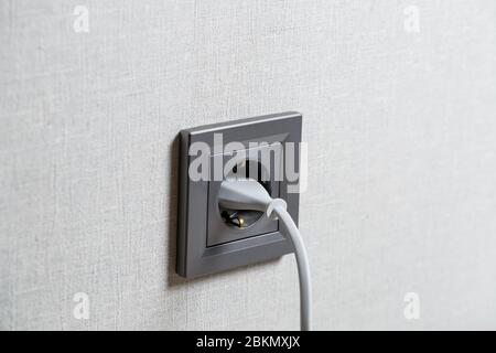 Eine graue Steckdose an der Wand, die ein Gerät mit einem weißen Kabel enthält. Laden über eine Steckdose Stockfoto