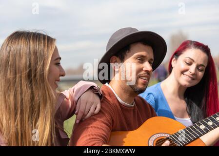 Ein hispanischer Junge in einem Hut spielt neben zwei kaukasischen Mädchen in einem Stadtpark die Gitarre Stockfoto