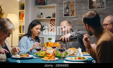 Ehemann gießt Rotwein zu ihrer schönen Frau beim Familienessen für mehrere Generationen. Stockfoto