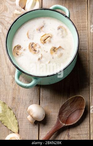 Draufsicht der türkisfarbenen Keramikpfanne mit Pilzcremesuppe, frischen Champignons, Löffel, Knoblauch und Lorbeer auf Holzhintergrund. Nahaufnahme. Stockfoto