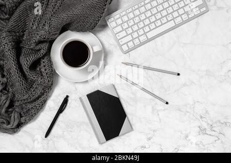 Marmorschreibtisch mit warmer Wolldecke oder Schal, Kaffeetasse, Tastatur, Notizblock, Stift und zwei Bleistiften in schwarz-weiß. Gemütlicher Arbeitsbereich im Home Office. Draufsicht Stockfoto