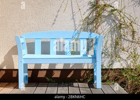 Schöne Holzgartenbank in hellblau lackiert, die an einem sonnigen Frühlingstag vor einer Hauswand auf einer Terrasse steht. Gesehen in Deutschland im April Stockfoto