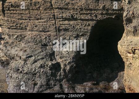 Dunkles vulkanisches Lavagestein an der Küste des Indischen Ozeans von Bali. Eine kleine Höhle, die von einer Brandung in eine Klippe ausgegraben wurde. Sonnenstrahlen beleuchten die raue wa Stockfoto