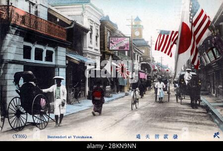 [ 1900er Japan - Japanese Shopping Street, Yokohama ] - Rikschas, ein Radfahrer und eine Pferdekutsche fahren unter Flaggen in Bentendori, Yokohama, Kanagawa Präfektur. Während der Meiji-Periode (1868-1912) war dies Yokohamas Premier Einkaufsstraße. Im Hintergrund ist der Uhrturm des Uhrenhändlers Kawakita Naozo Shaten zu sehen. Es war das wichtigste Wahrzeichen der Straße. Vintage-Postkarte des 20. Jahrhunderts. Stockfoto