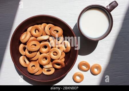 Milch in einem braunen Keramiktassen und kleine trockene Bagels für Kinder auf weißem Holzhintergrund. Erste feste Fingernahrung für Baby. Gesunde Mahlzeit Stockfoto
