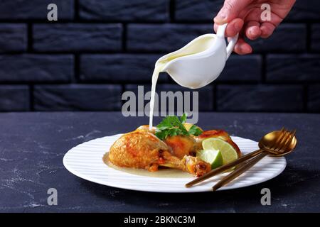 Frau Hand gießt tropfende Kokosnuss-Sauce auf gebratene Hähnchenschenkel serviert mit Babykartoffeln auf einem weißen Teller, horizontale Ansicht von oben Stockfoto