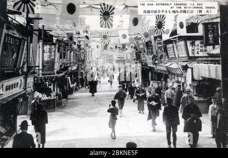 [ Japan der 1920er Jahre - Osaka Entertainment District ] - Menschen gehen unter Flaggen in Sennichimae, Osaka. Seit der Edo-Periode (1603-1868) war Sennichimae zusammen mit Dotonbori Osaka das Hauptunterhaltungs-Viertel. Vintage-Postkarte des 20. Jahrhunderts. Stockfoto