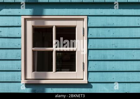Ein kleines Fenster eines kleinen Hauses mit einem weißen Rahmen und hellblau gefärbte harte Holzfassade, bestehend aus Paneelen an einem sonnigen Tag. Horizontale Aufnahme Stockfoto