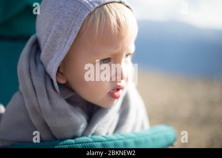 Ein blonder Junge sitzt in einem Kinderwagen, der in eine Decke gehüllt ist.EIN blonder Junge sitzt in einem Kinderwagen, der in eine Decke gehüllt ist. Stockfoto