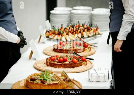 Kellner, deren Gesichter nicht zu sehen sind, stehen neben dem Banketttisch, auf dem liegende Kuchen mit Kirschtomaten und grünen Keimen auf runden Holztabletts sich befinden Stockfoto