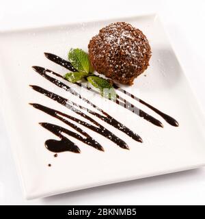 Ein rundes Stück Kuchen, mit Puderzucker bestreut und mit Minze und geschmolzener Schokolade auf einem weißen quadratischen Teller dekoriert Stockfoto