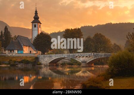 Kirche des heiligen Johannes des Täufers und mittelalterliche Steinbrücke am Bohinjer See, Slowenien. Das Bild wurde am frühen Morgen des august aufgenommen. Stockfoto