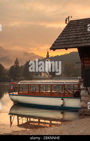 Elektrisches Motorboot für die Besichtigung des Bohinjer Sees in Slowenien, Kirche des heiligen Johannes des Täufers und alte Steinbrücke im Hintergrund Stockfoto