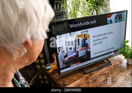 Ältere Frau, die die Nachrichten mit der Überschrift "weltweite Virustodesfälle passieren 250,000" und einem Bild von einem Patienten, der in einen Krankenwagen gehoben wird, beobachtet. Stockfoto