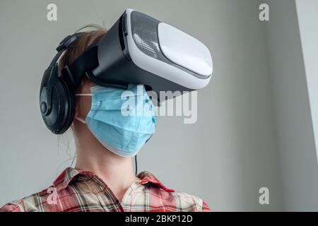 Porträt einer Frau, die eine medizinische Gesichtsmaske, ein Virtual Reality Headset trägt und sich zu Hause umsieht - niedrige Blickwinkel. VR, Selbstisolierung, Prävention Stockfoto