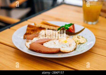 Hilfreiches appetitliches Frühstück auf einem weißen Teller. Rührei mit Gemüse und Croutons in einem Café. Frühstück im Café: Rührei, Würstchen Stockfoto