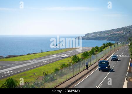 Mann beobachtet Flugzeug, das von der Start- und Landebahn des internationalen Flughafens Funchal abfährt, landschaftlich schönen Blick aufs Meer und die Stadt im Hintergrund, Autos auf der Autobahn, Madeira, Stockfoto