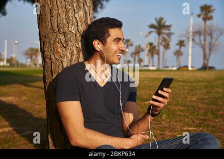 Ein Mann, der sich in einem Park an einen Baumstamm lehnt und über Kopfhörer etwas auf seinem Mobiltelefon hört. Stockfoto