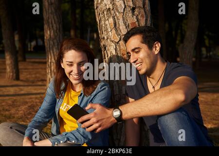 Ein Paar, das an einem Baumstamm sitzt und ein Mobiltelefon anlächelt. Stockfoto
