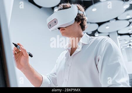 Junger Mann mit Virtual Reality Brille Schreiben auf Flipchart während der Online-Präsentation vor Ort ohne Zuschauer. Soziale Distanz. Bleib Zu Hause Stockfoto